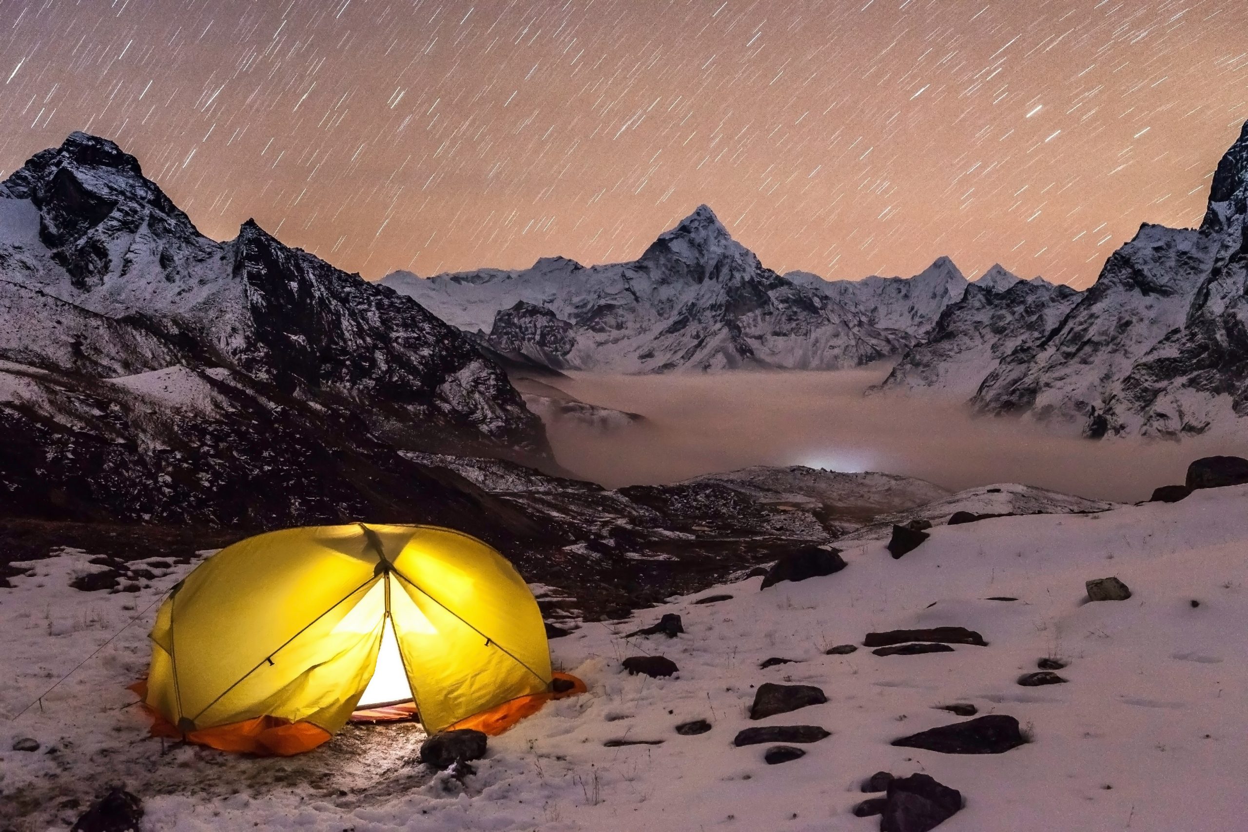 Illuminated yellow tent set against a snow-covered Himalayan landscape under a sky full of star trails, showcasing the serene experience of camping trekking in Nepal.